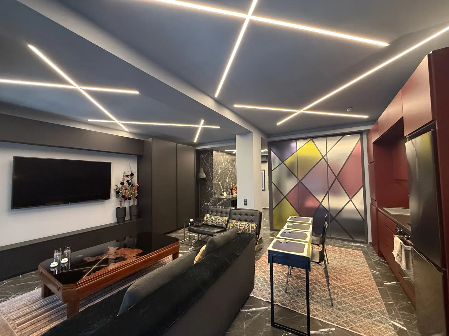 A modern apartment interior with black marble floors, a black sofa, and a kitchen with red cabinets. Linear lights crisscross the ceiling.