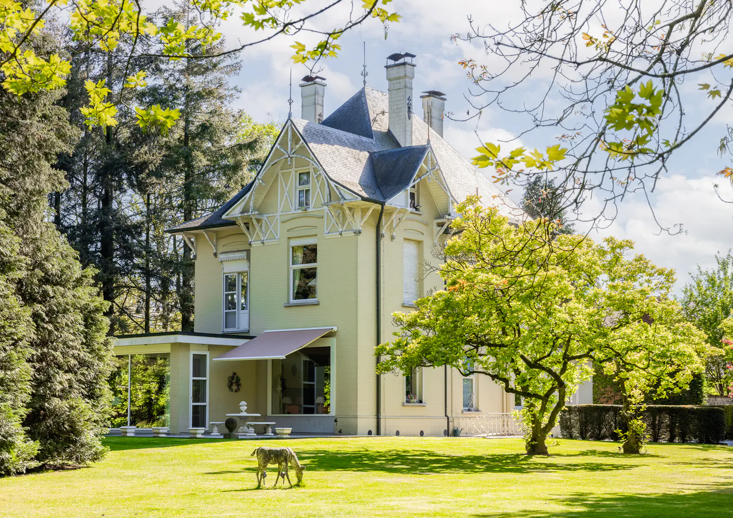 A pale yellow two-story house with a gray roof sits on a green lawn with trees. A deer statue grazes in the yard.