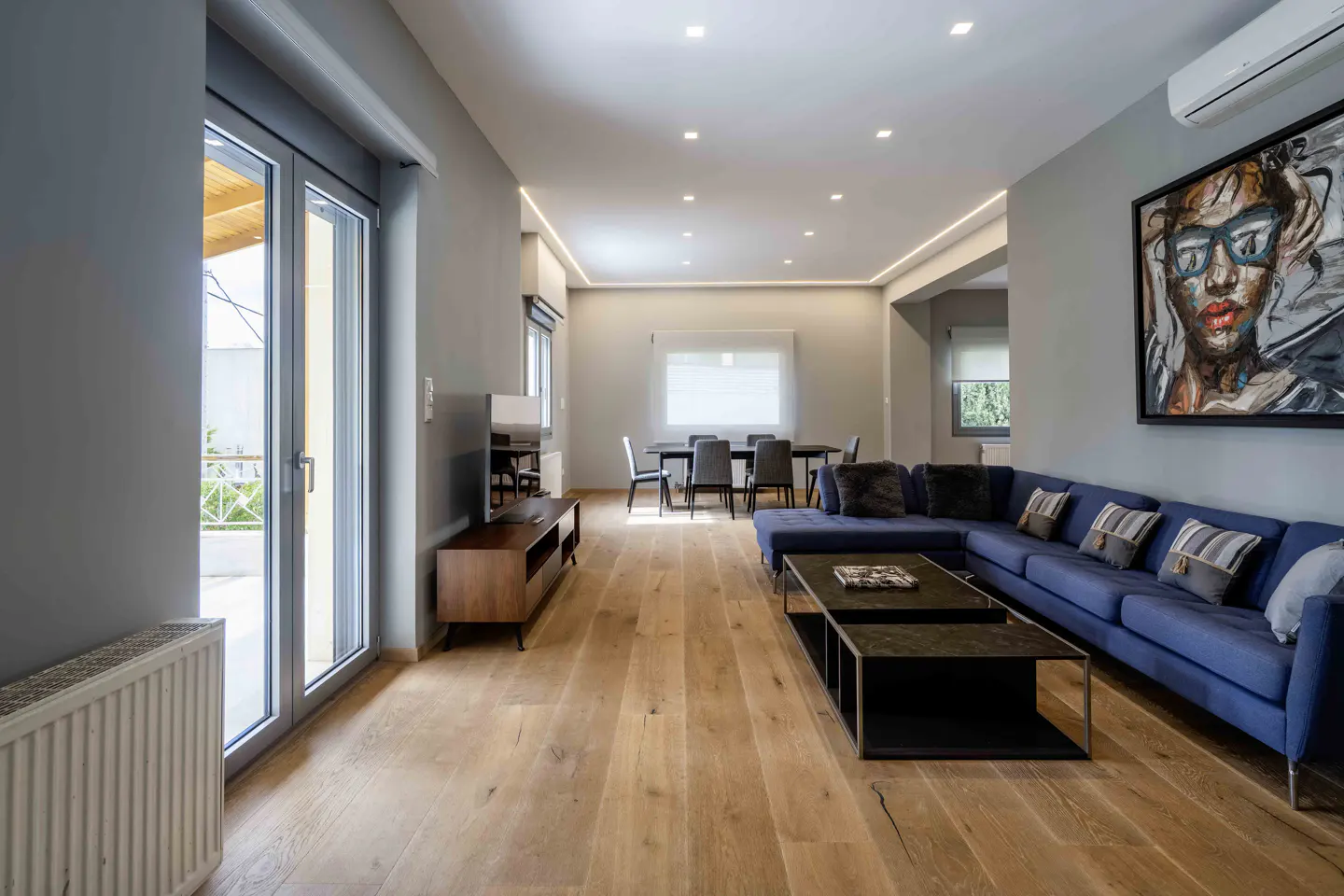 Modern living room with blue sectional sofa, wooden floors, coffee tables, and dining area in the background.