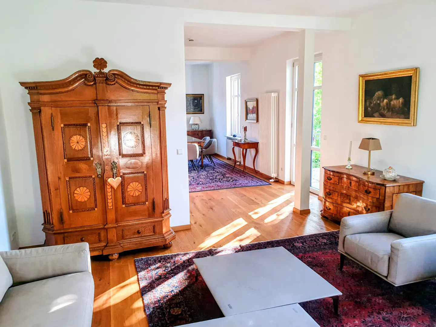 Bright living room with hardwood floors, antique wooden armoire, and gray sofas. A dining room is visible through an archway.