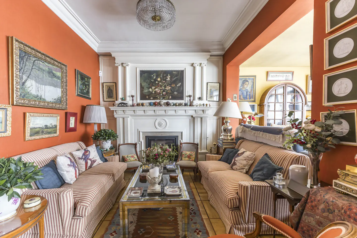 A living room with orange walls, striped sofas, a white fireplace, and many framed pictures. A chandelier hangs from the ceiling.