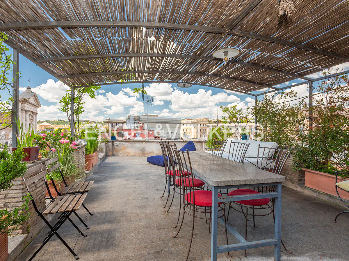 Rooftop terrace with a stone dining table, red-cushioned chairs, and a bamboo pergola overlooking a historic city skyline.