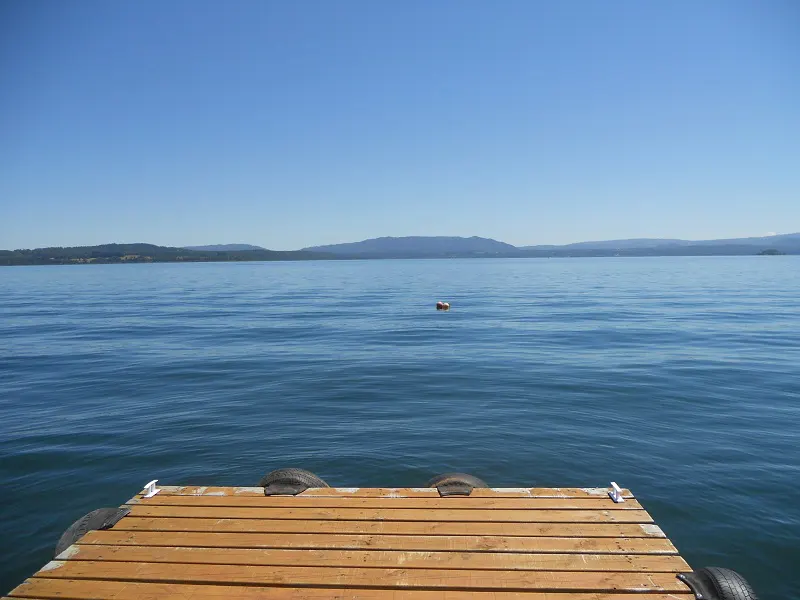 A wooden dock extends into a calm, blue lake under a clear sky. Mountains are visible in the distance.