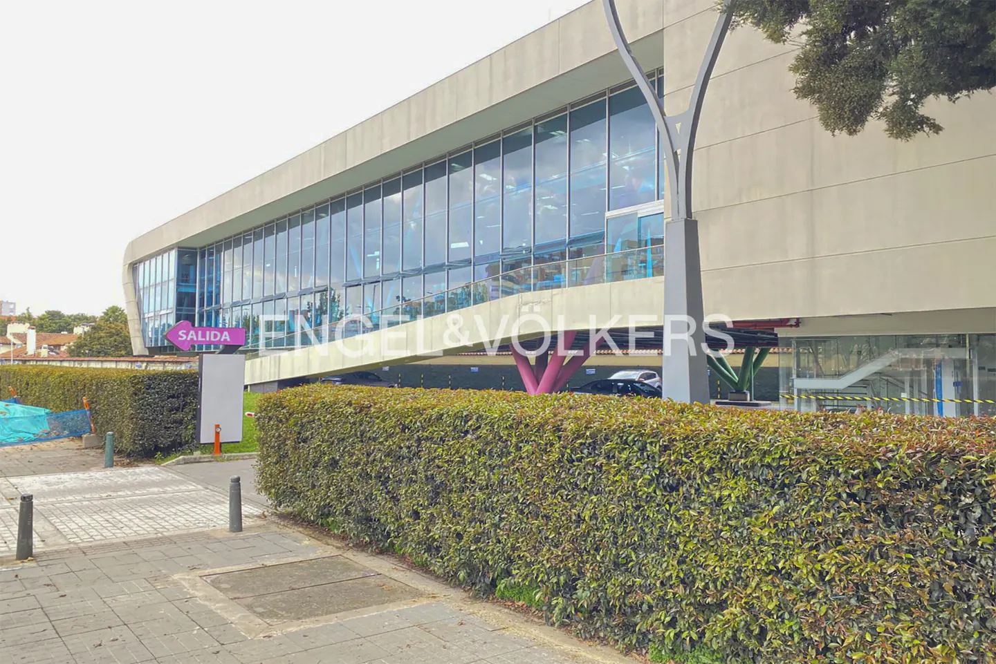 Exterior view of a modern, multi-story building with large windows, green hedges, and a pink "Salida" (Exit) sign.