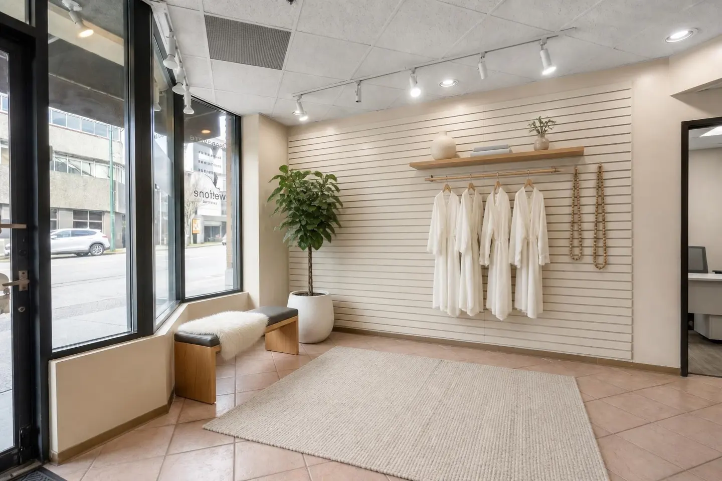 Interior of a boutique with white robes hanging on a wall, a plant, and a bench near a window.