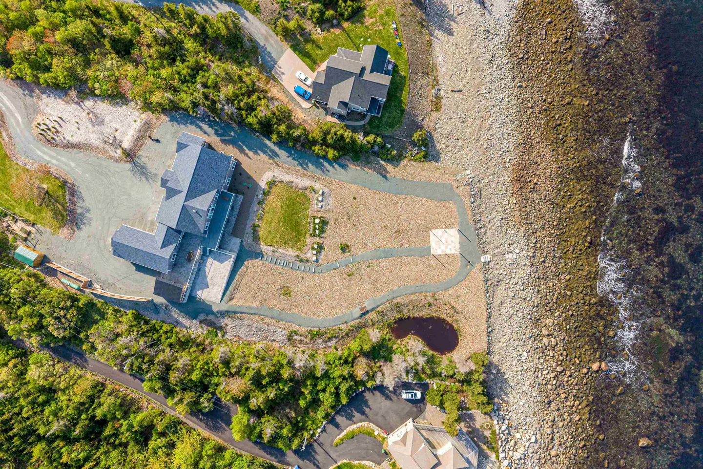 Aerial view of a coastal property with a grey house, gravel paths, a small pond, and lush trees bordering the rocky shore.