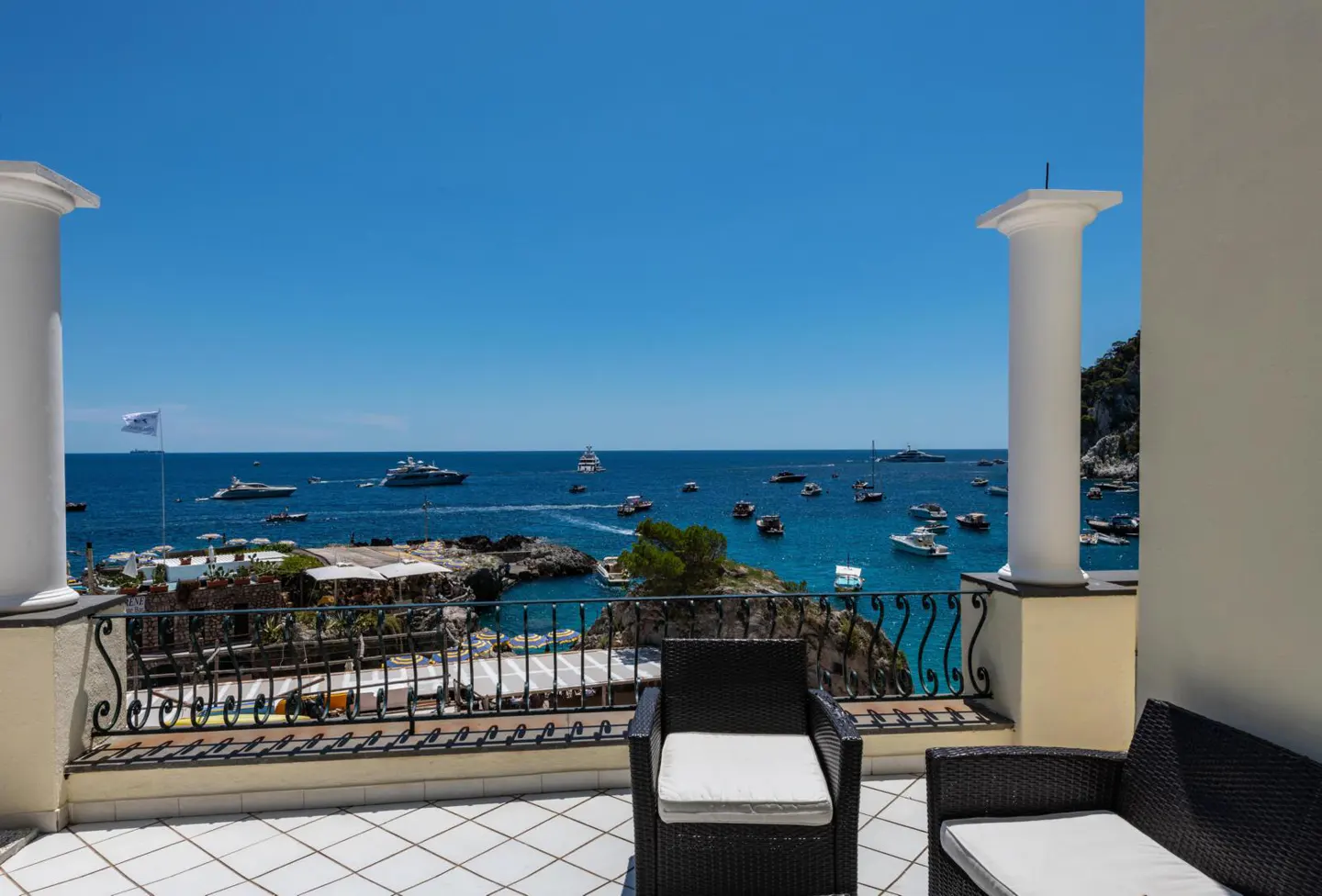 Balcony view with black wicker chairs overlooking a blue ocean filled with boats under a clear blue sky.