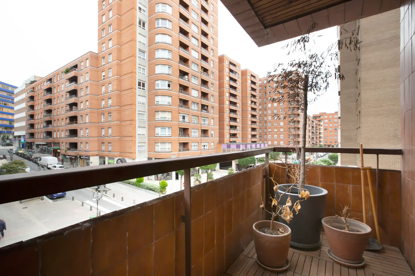 View from a balcony with brown railings and potted plants, overlooking a city street with tall brick apartment buildings.