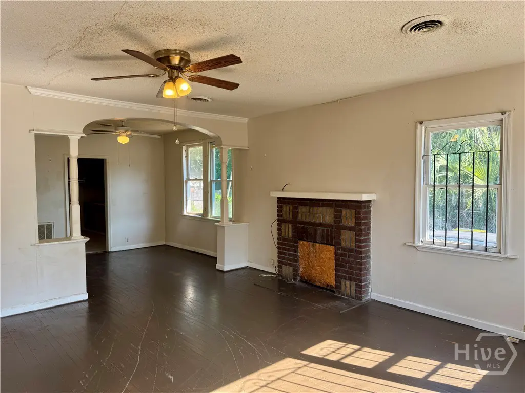 Living room with dark wood floors, beige walls, a brick fireplace, ceiling fan, and a window with security bars.