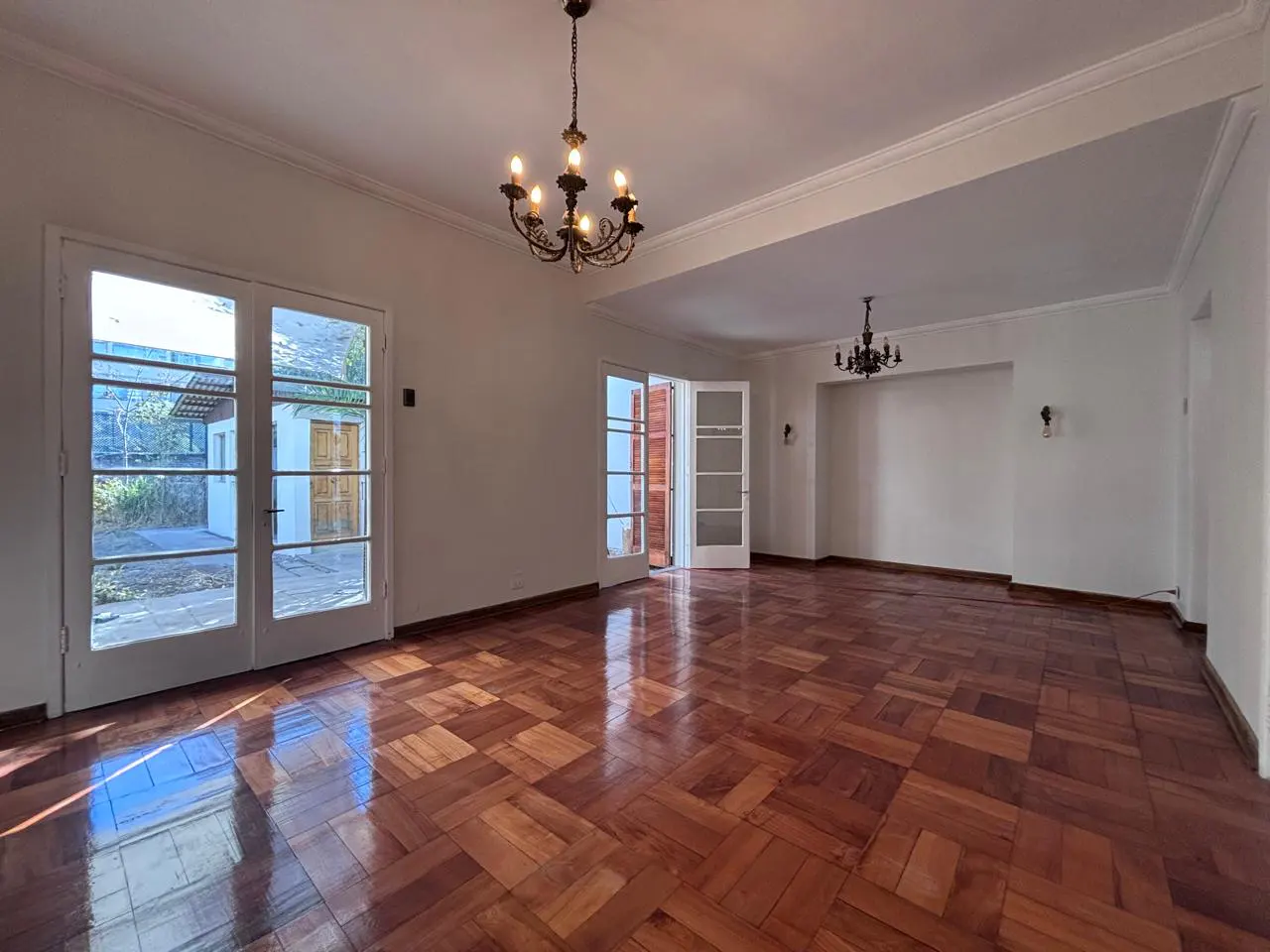 An empty living room with parquet floors, white walls, and two chandeliers. French doors lead to a patio.