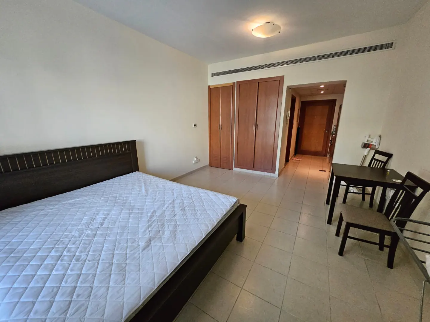 Studio apartment with a dark wood bed, white quilt, dining table with chairs, and wood wardrobes against cream walls.