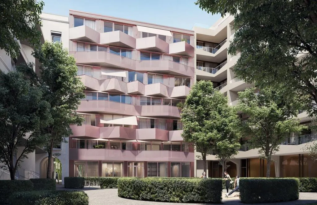 Exterior view of a pink apartment building with geometric balconies, surrounded by green trees and manicured hedges.