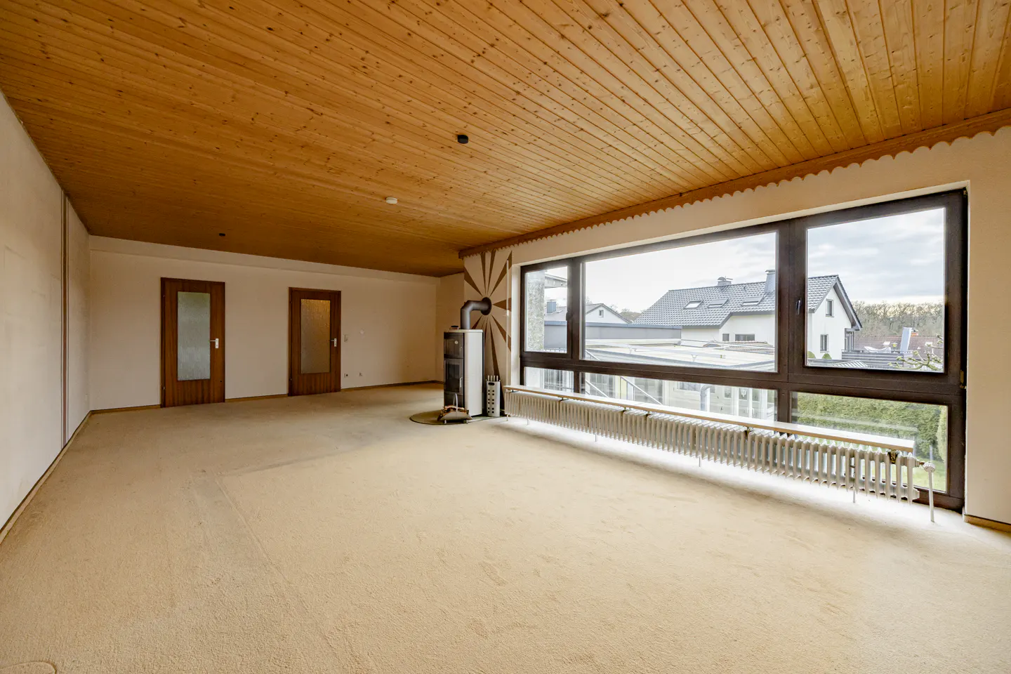 Empty room with beige carpet, wood-paneled ceiling, two wooden doors, a fireplace, and a large window overlooking houses.