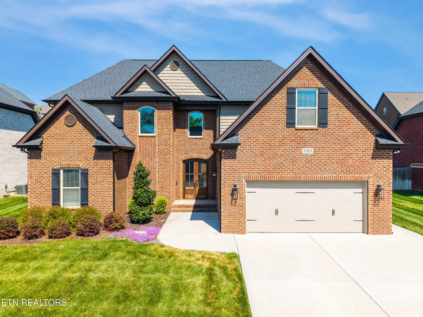 Two-story brick house with a gray roof, black shutters, and a two-car garage on a sunny day.