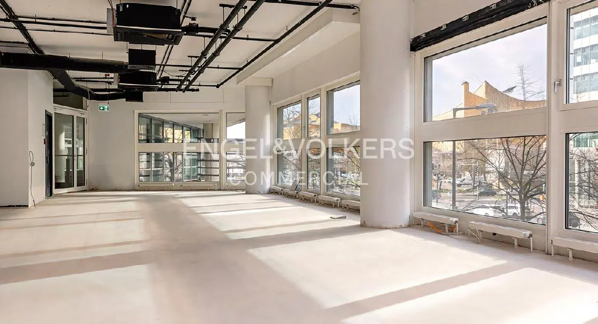 Bright, empty commercial space with white walls, large windows, and black ceiling pipes. Sunlight streams across the floor.