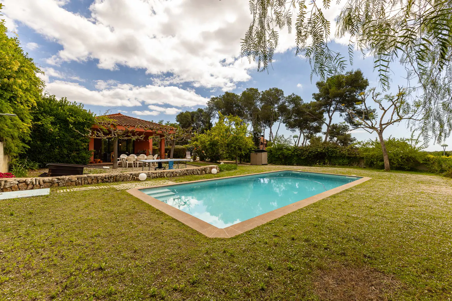 A backyard with a triangular pool, green grass, and a red-roofed pergola with a table and chairs.