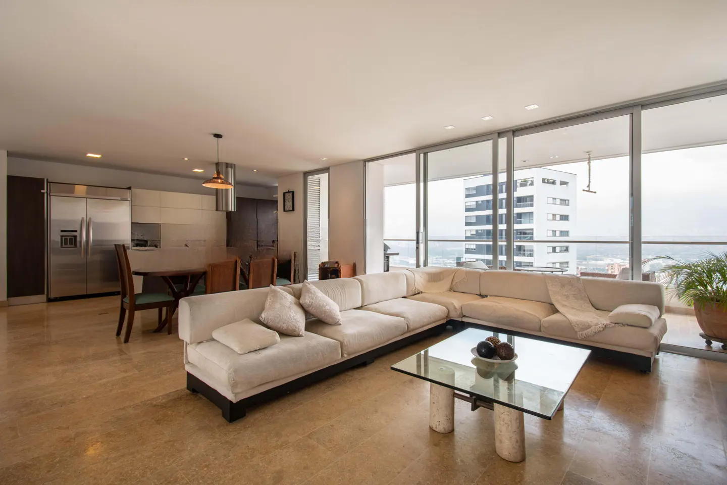 Modern open-plan living room with a large white sectional sofa, glass coffee table, and kitchen with stainless appliances.