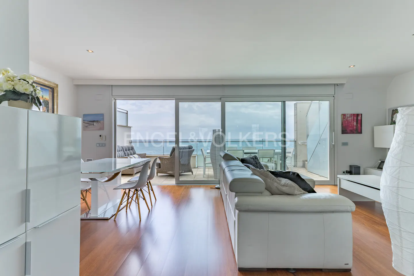 Bright, modern living room with white sofa, dining table, and ocean view through sliding glass doors. Hardwood floors and white walls.