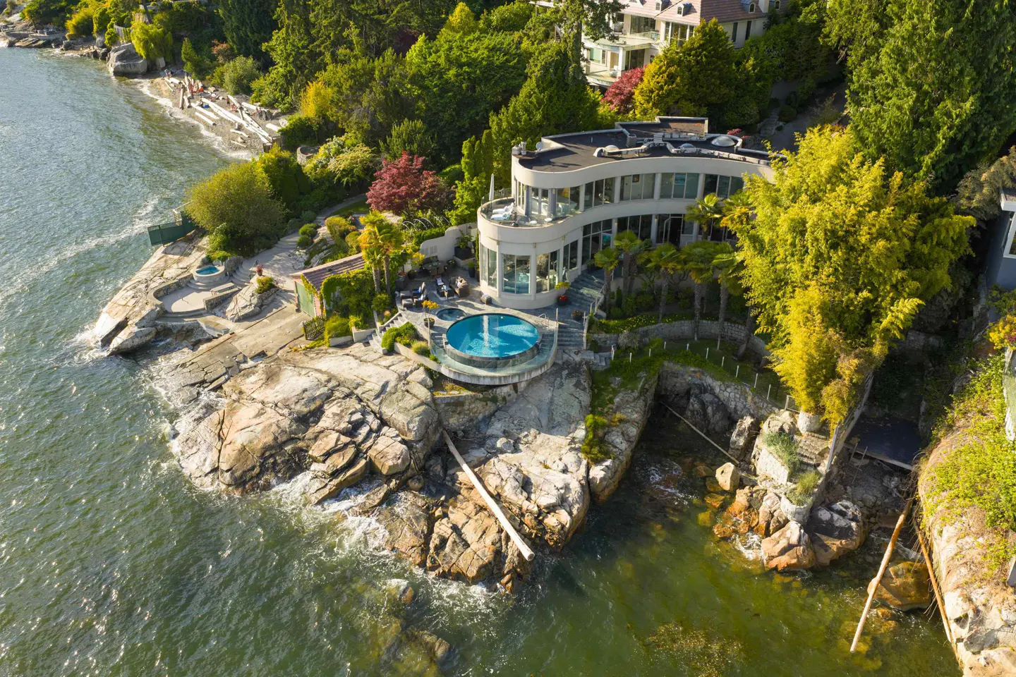 Aerial view of a modern curved waterfront home with a circular pool, nestled on a rocky coastline surrounded by lush trees.