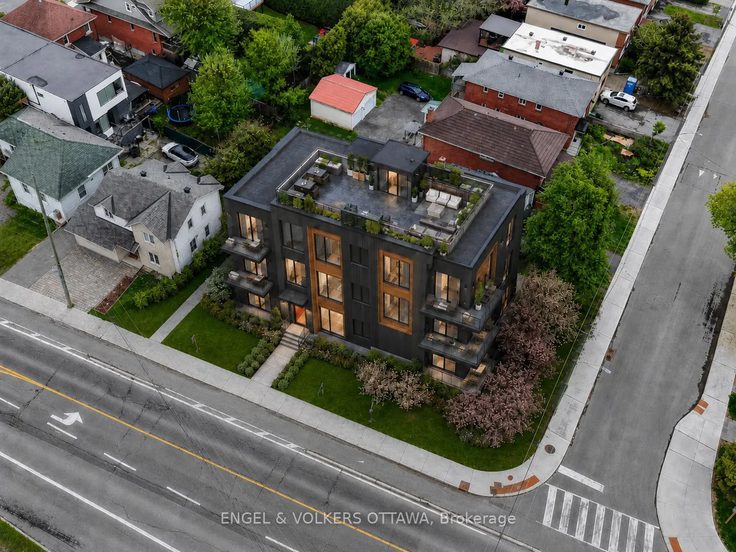 Aerial view of a modern black apartment building with a rooftop terrace, surrounded by residential homes and a city street.