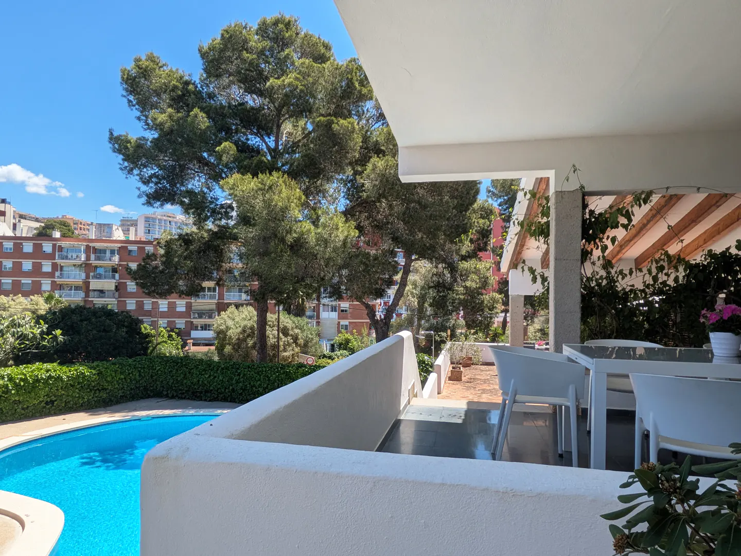 Balcony view of a blue pool, trees, and buildings under a clear sky. White table and chairs are on the patio.