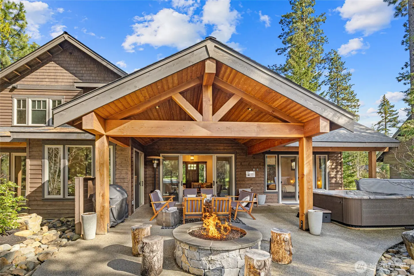 Outdoor patio with a stone fire pit, wood chairs, and a hot tub. The house is brown with a wood beam roof. Tall trees are in the background.