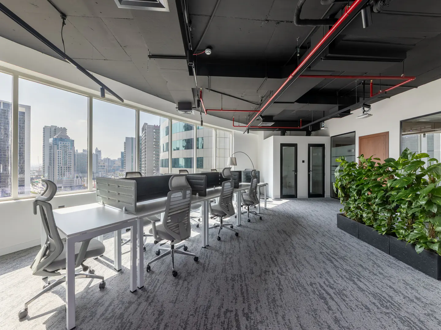 Bright office space with white desks, gray chairs, and a city view through large windows. Black ceiling with exposed pipes and a planter with green plants.