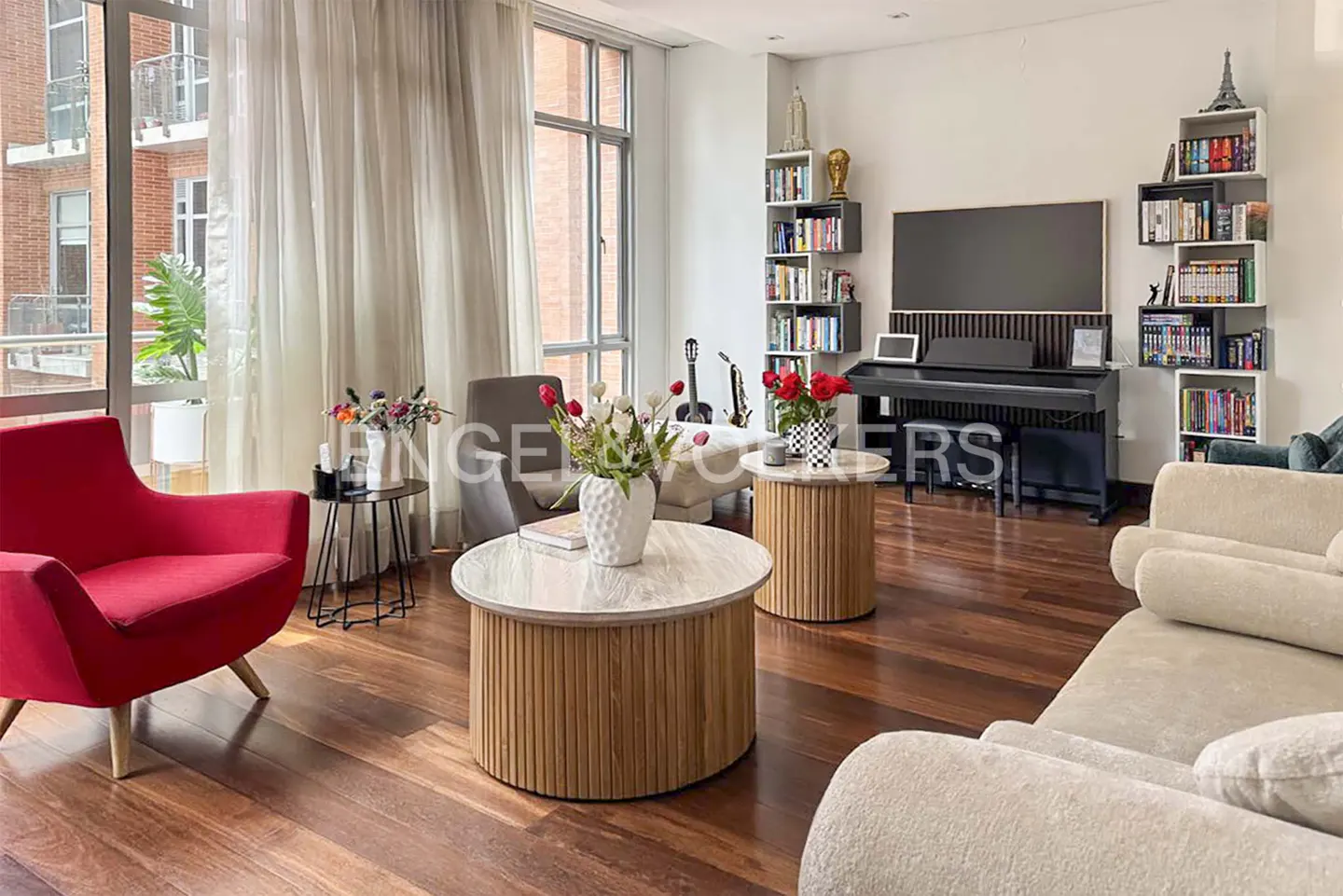 Bright living room with a red armchair, beige sofa, two round wooden coffee tables, a piano, and floor-to-ceiling windows.