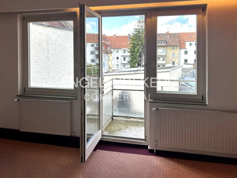 A room with a window and open balcony door, showing a view of buildings and trees outside. The room has a radiator and patterned carpet.