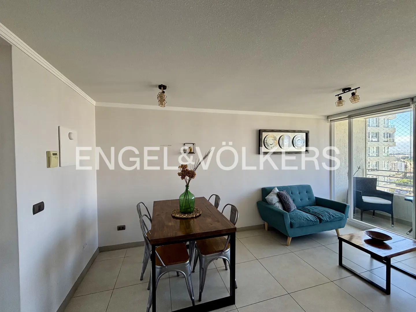 Living room with a dark wood table, teal sofa, and balcony with a view. Engel & Völkers logo is visible.