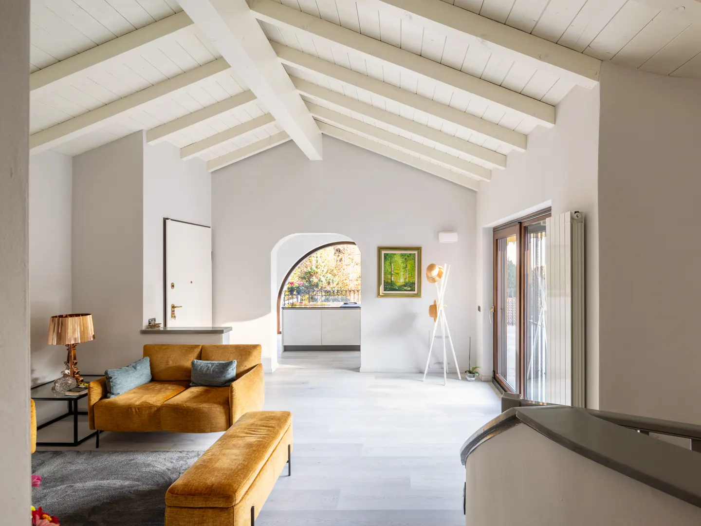 Bright, airy living room with vaulted ceiling, white beams, and gray walls. A mustard-colored sofa and bench sit on a gray rug. Arched doorway leads to kitchen.