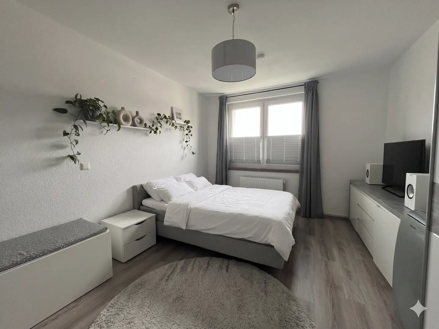 Bright bedroom with a gray bed, white linens, and a round rug. A shelf with plants and vases is on the wall. A TV and speakers sit on a dresser.
