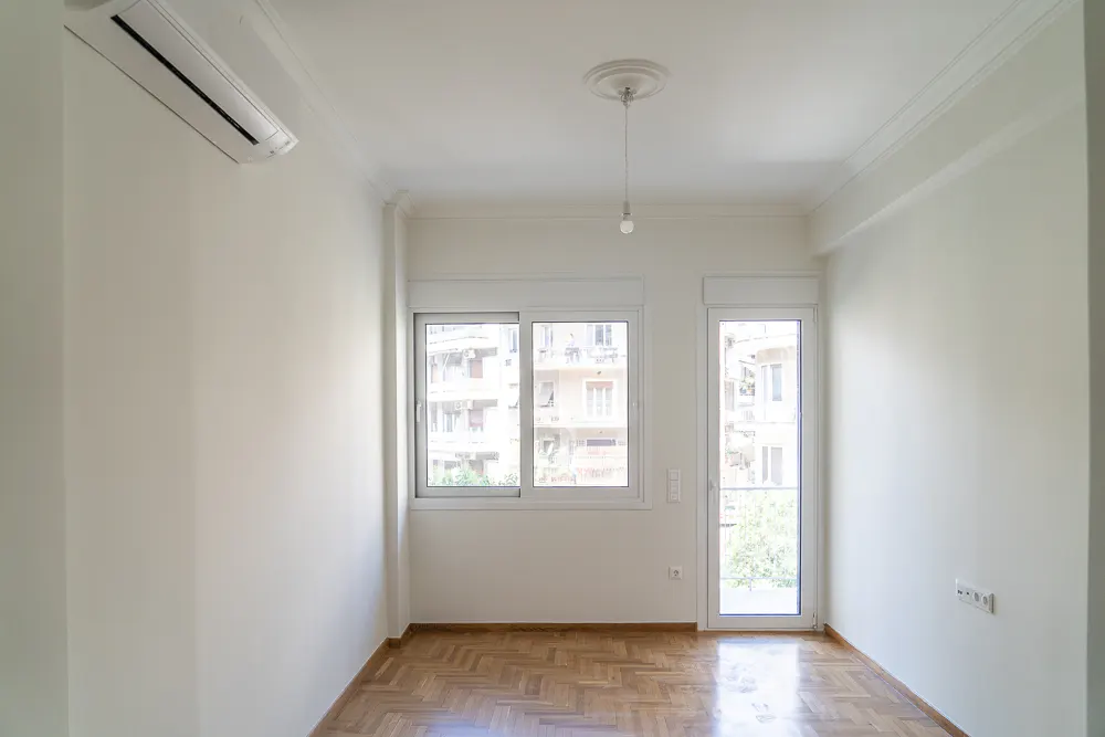 Bright, empty room with parquet floor, white walls, and a window. An air conditioner is mounted high on the wall.