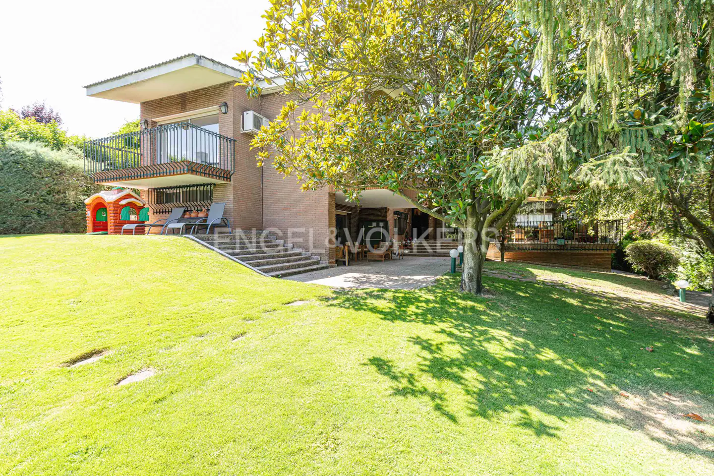 Brick house with a balcony, a green lawn, and a large tree in the front yard. A playhouse and chairs are on the lawn.