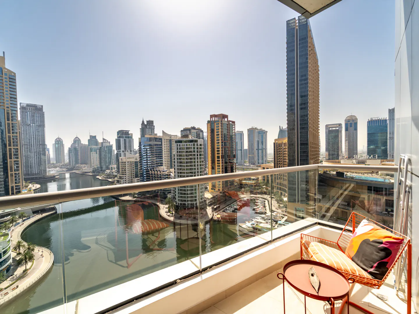 Balcony view of Dubai Marina skyscrapers and canal, featuring a small orange table and chair with a patterned cushion.