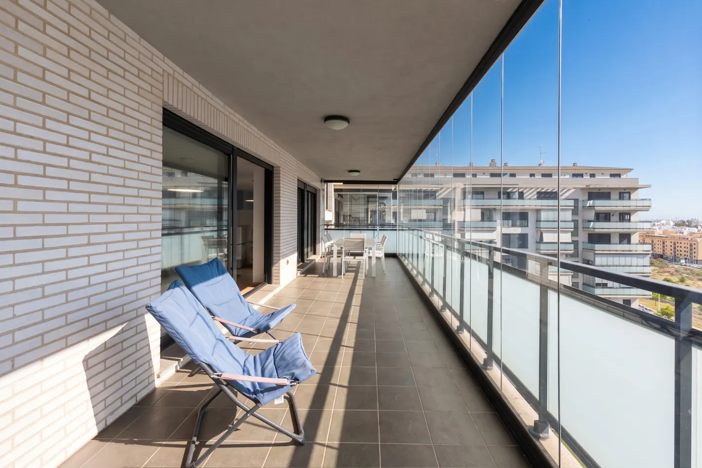 Spacious balcony with tiled floor, two blue folding chairs, white brick wall, and glass railings overlooking city buildings.