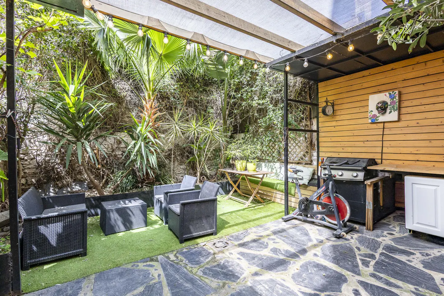 Outdoor patio with black wicker furniture, exercise bike, grill, and lush green plants. String lights hang above.