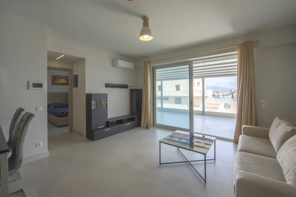 Bright living room with a beige sofa, glass coffee table, and dark entertainment center. Sliding glass doors lead to a balcony with a city view.