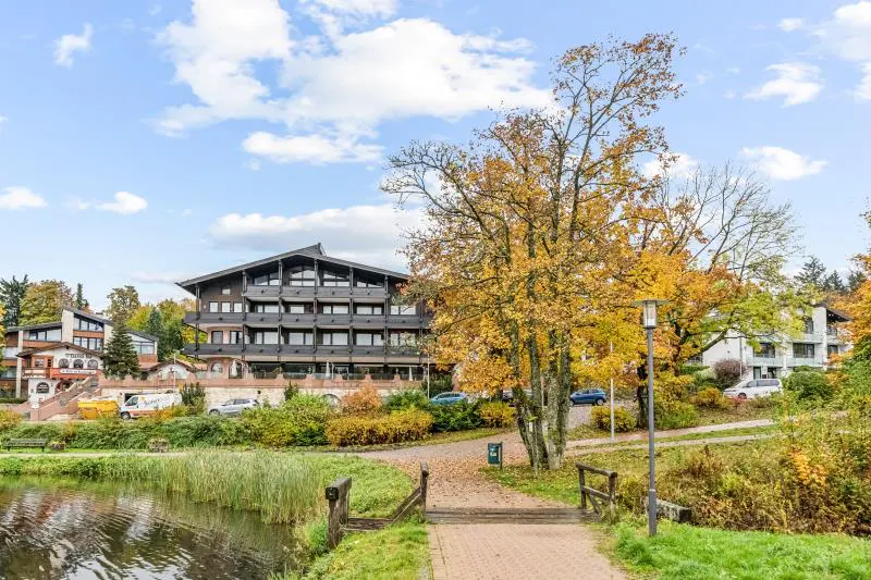 Exterior view of a multi-story hotel with dark wood trim, surrounded by trees with autumn foliage and a pond in the foreground.
