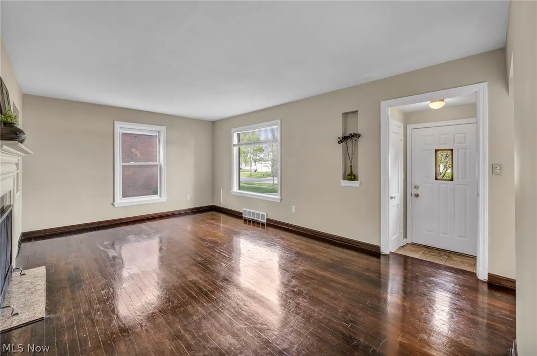 A bright, empty living room with dark wood floors, beige walls, and white trim. Two windows let in natural light. A white front door is visible.